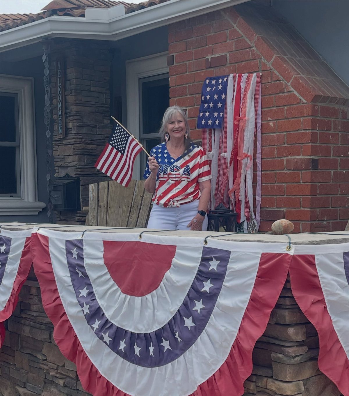 Woman holding an American flag on a porch decorated with patriotic bunting and streamers.