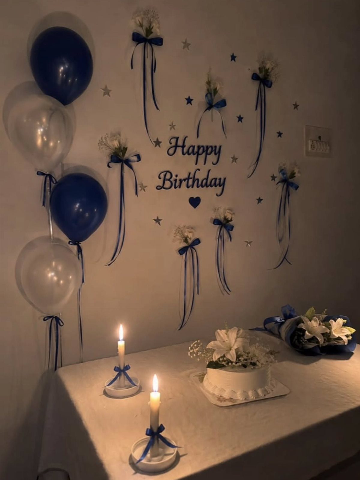 Birthday decorations with blue balloons, a white cake, and ribbons on a wall.