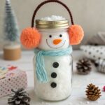 Mason jar snowman filled with white snow, wearing a blue scarf and orange earmuffs.