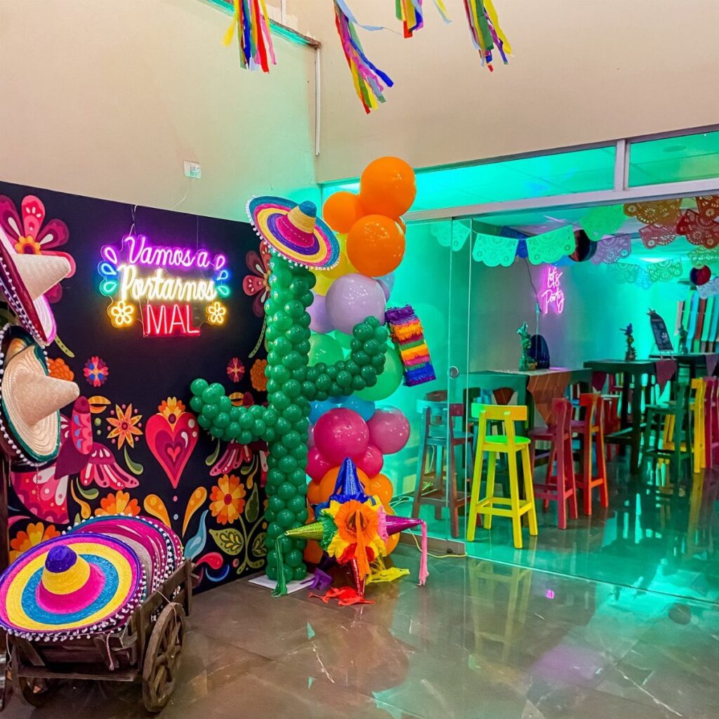 Mexican party decorations featuring a cactus balloon sculpture, colorful stools, and floral backdrop.